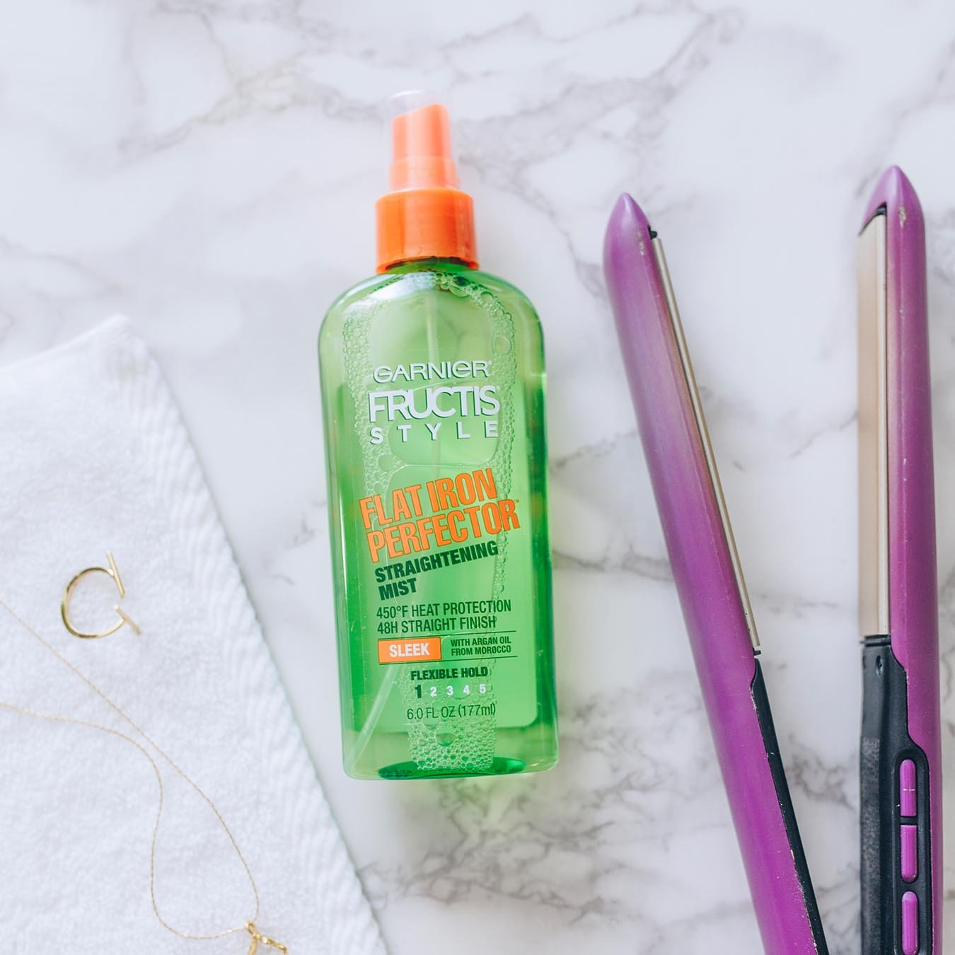 Garnier Fructis Style Flat Iron Perfector Straightening Mist on white marble next to purple hair straighteners and a gold earring and necklace on a white wash cloth.